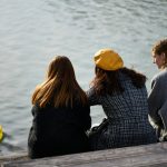 Three women sit by the water, observing a duck.